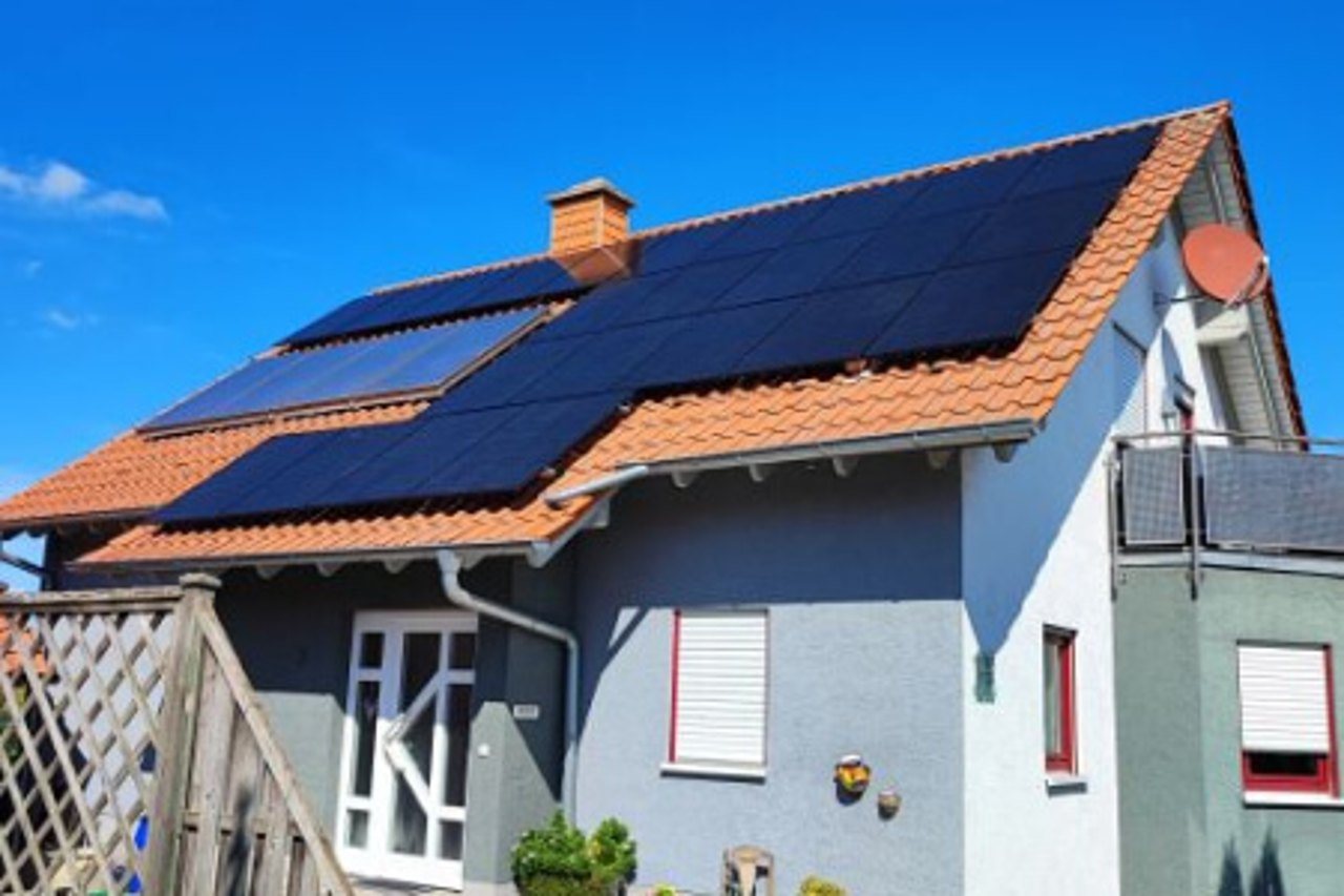 Graues Haus mit rotem Ziegeldach und schwarzen Solarmodulen unter blauem Himmel.